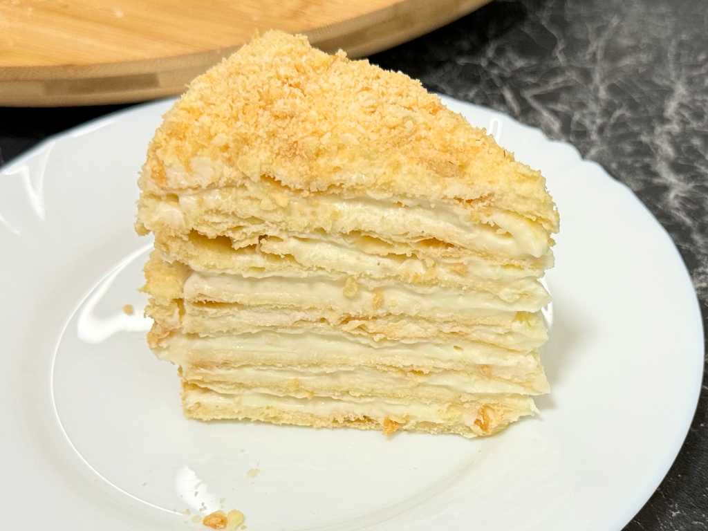 Slice of Napoleon Cake on a white plate, showing layers of flaky puff pastry and creamy custard topped with fine crumbs.