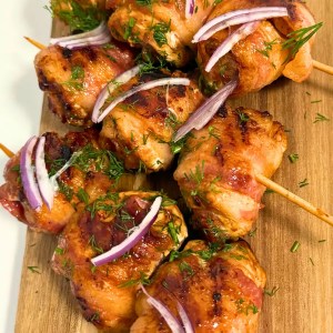 Juicy Bacon Wrapped Chicken Skewers topped with fresh dill and red onion slices, served with tomato dipping sauce.