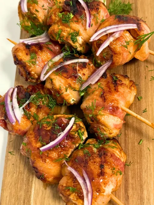 Juicy Bacon Wrapped Chicken Skewers topped with fresh dill and red onion slices, served with tomato dipping sauce.