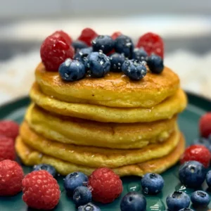 Stack of golden fluffy pancakes topped with raspberries and blueberries, served on a green plate