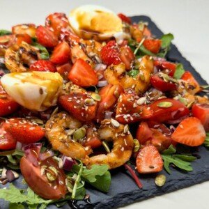 Colorful shrimp salad with strawberries, cherry tomatoes, mixed greens, seeds, and balsamic glaze topped with soft-boiled egg.