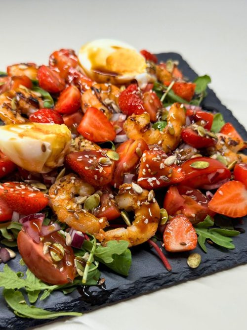 Colorful shrimp salad with strawberries, cherry tomatoes, mixed greens, seeds, and balsamic glaze topped with soft-boiled egg.