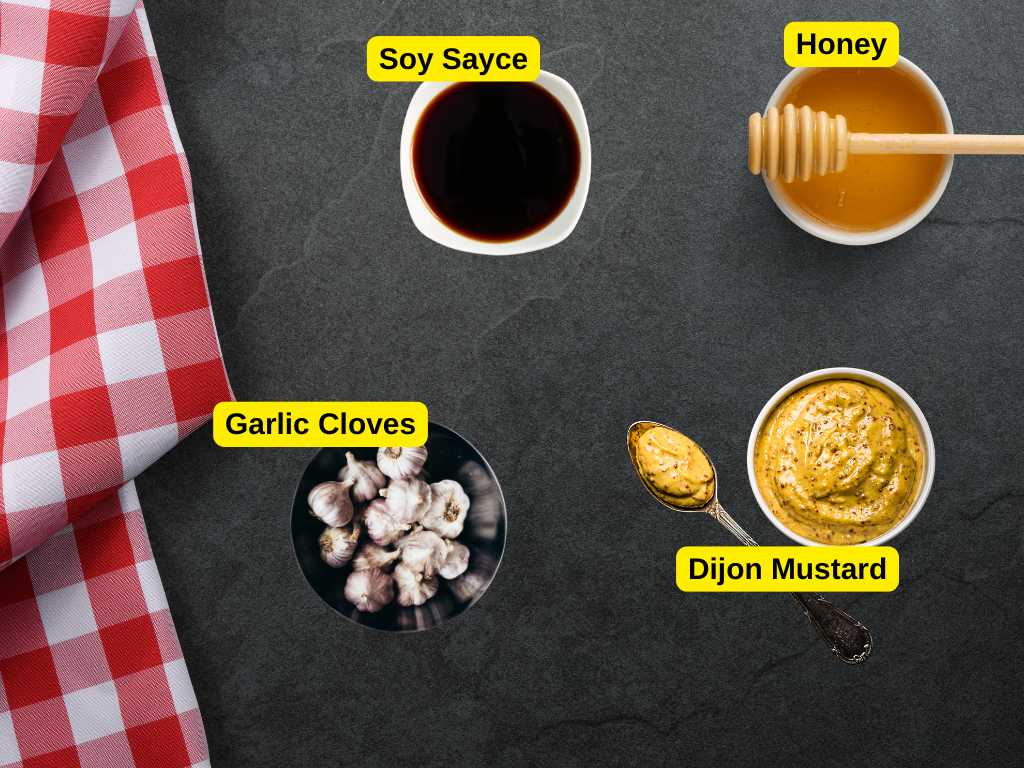 Overhead view of honey, soy sauce, Dijon mustard, and garlic cloves arranged on a dark surface with a red checkered cloth.