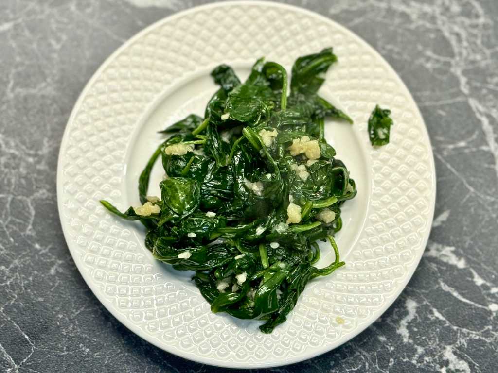 Cooked spinach sautéed with minced garlic, served on a white textured plate over a dark marble surface.