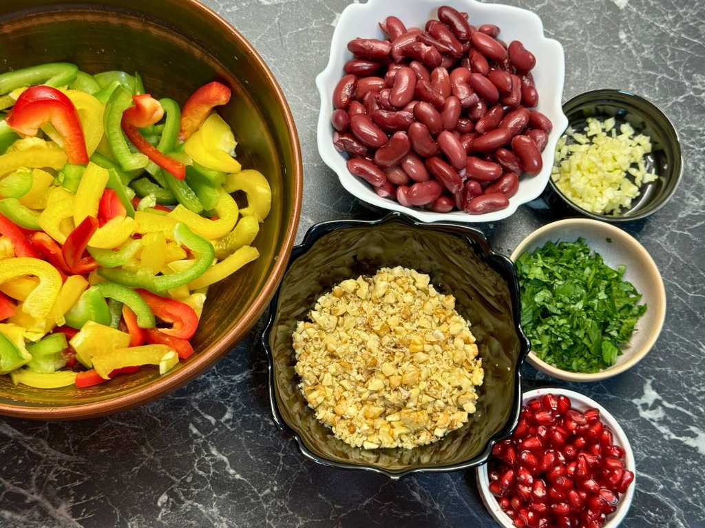 Prepared ingredients for Tbilisi Salad, including sliced bell peppers, cooked beans, chopped garlic, fresh parsley, chopped walnuts, and pomegranate seeds.