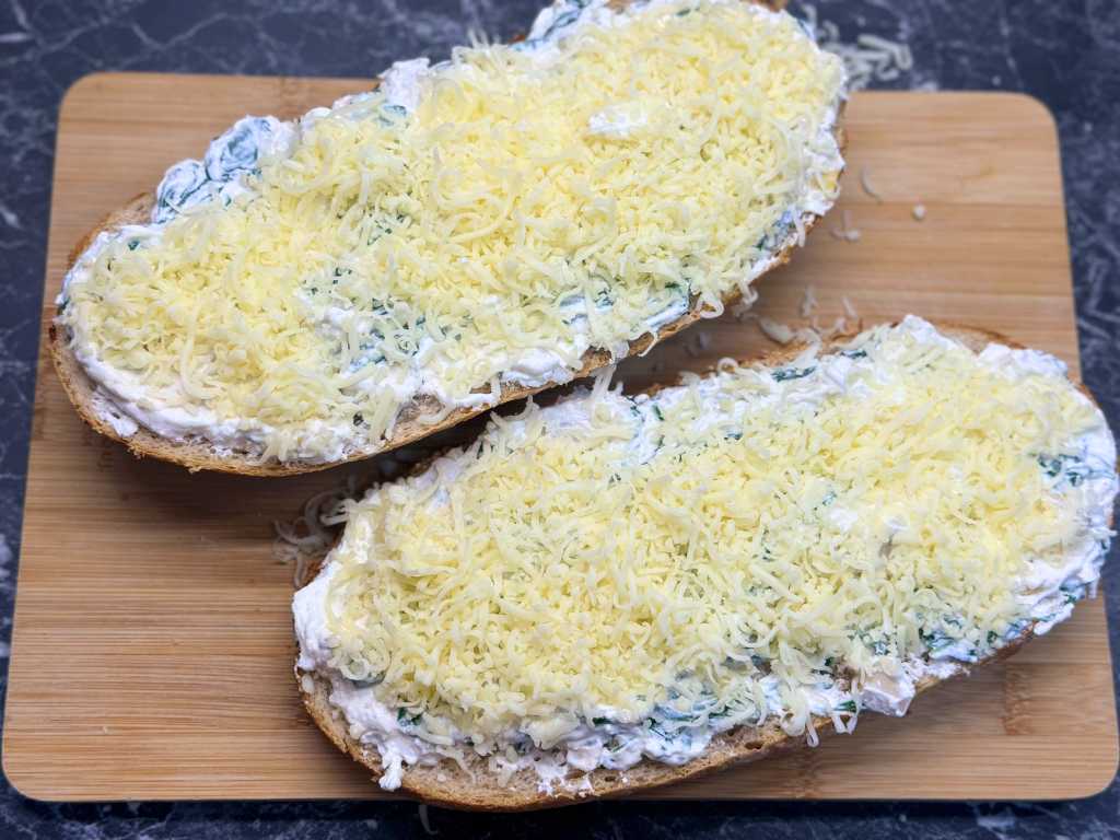 Sourdough bread halves spread with creamy spinach artichoke mixture and topped with shredded mozzarella, ready for baking.