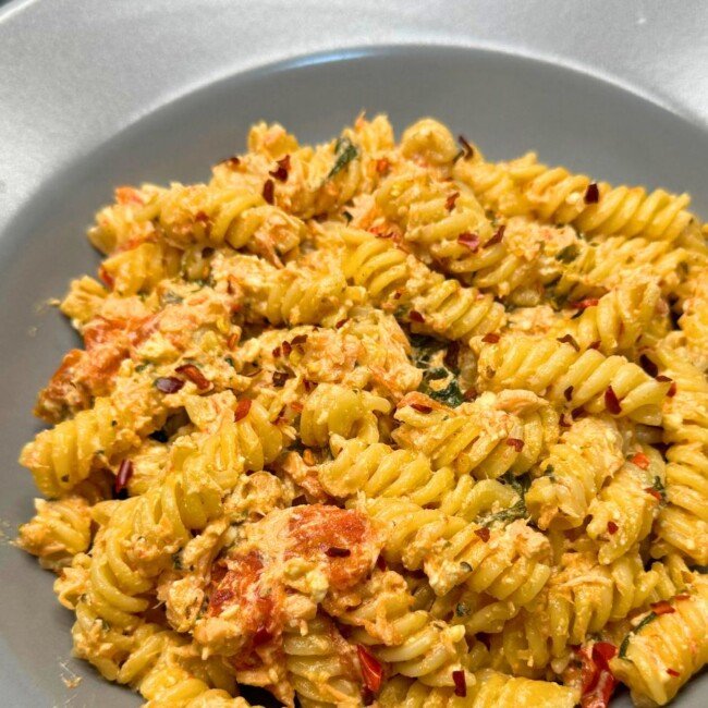 Creamy salmon and feta pasta with fusilli, roasted cherry tomatoes, and spinach, topped with red pepper flakes in a gray bowl.