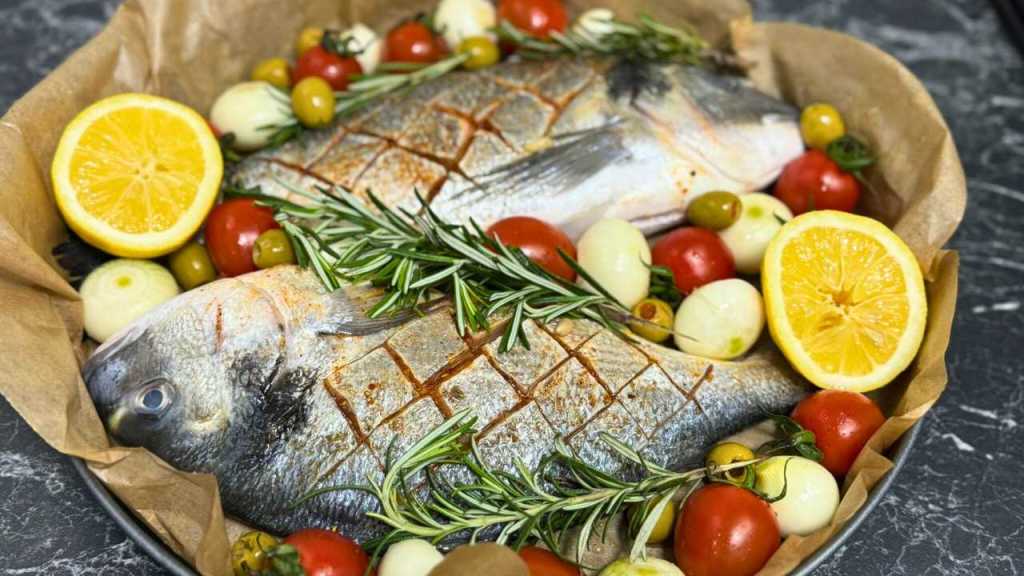 Two whole dorada fish in a roasting pan, surrounded by cherry tomatoes, pearl onions, green olives, lemon halves, and fresh rosemary sprigs.