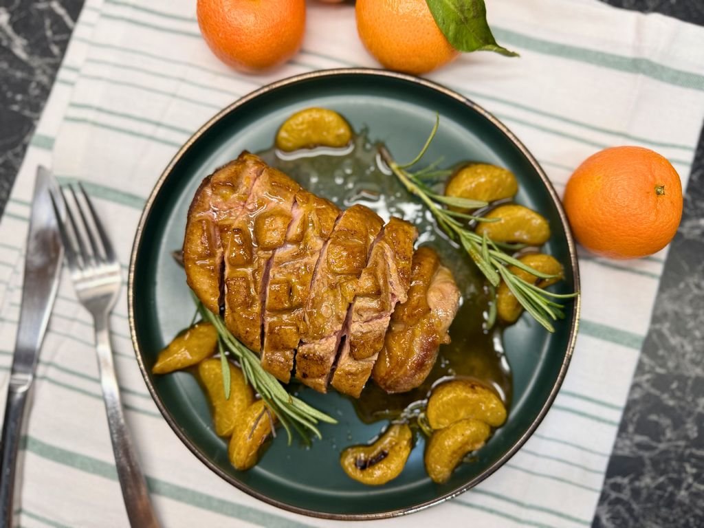 Sliced honey orange glazed duck breast served on a dark green plate with caramelized clementine segments and rosemary sprigs, styled on a striped linen napkin with whole clementines beside the plate.