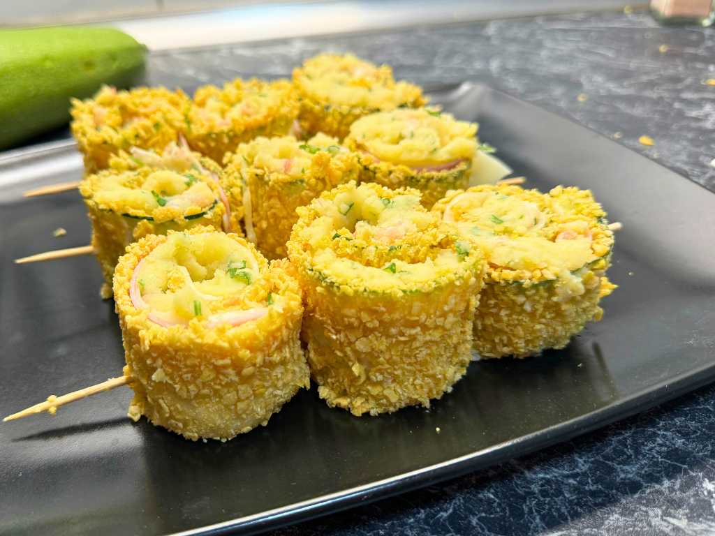 Unbaked zucchini roll skewers with mashed potato, ham, and cheese filling, coated in golden breadcrumbs and arranged on a black plate