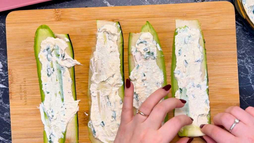 Four zucchini slices laid flat on a wooden board, each spread with a layer of ricotta filling. Hands are preparing to roll one of the slices.