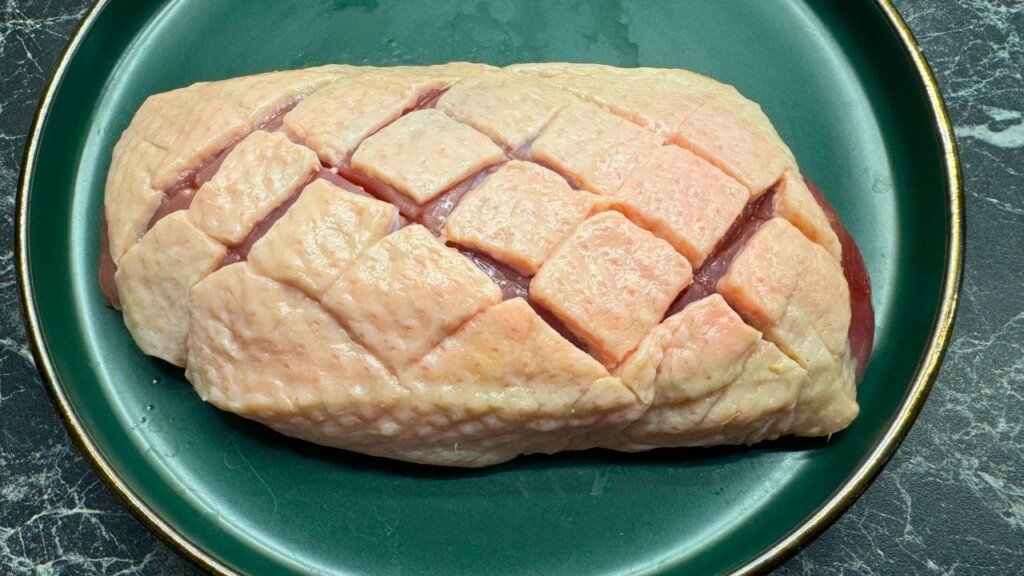 Raw duck breast with crosshatch scoring on the fat side, prepared for pan-searing, displayed on a green plate—ideal for crispy skin in duck breast recipes.