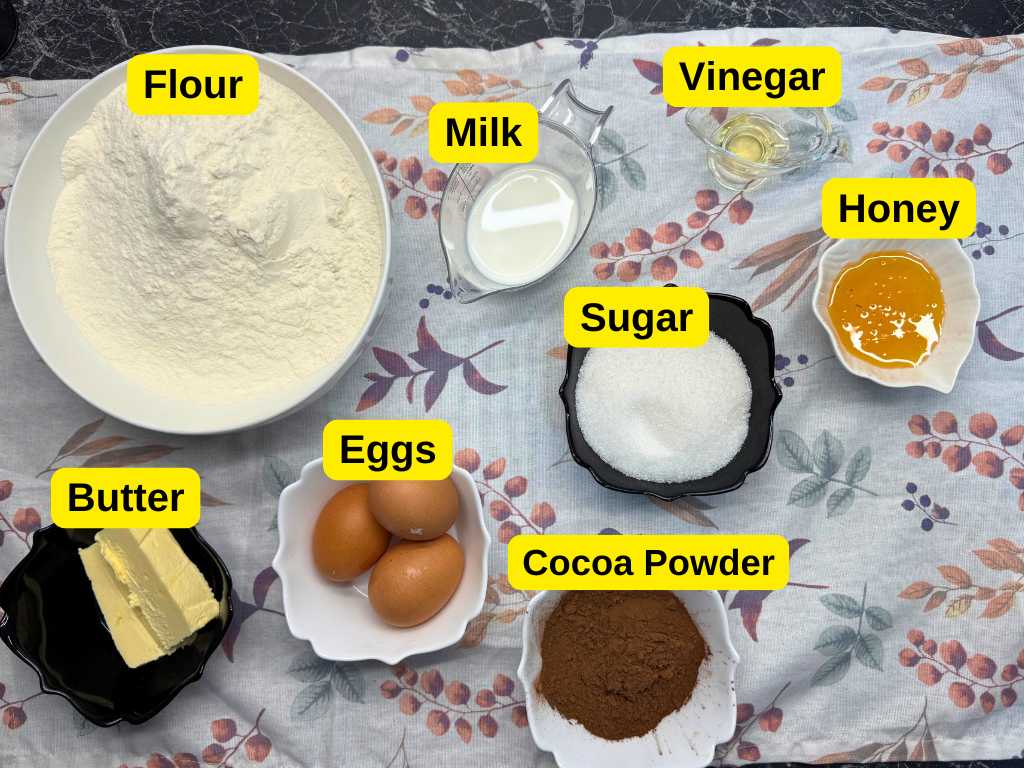 Top view of Spartak Cake ingredients labeled: flour, milk, vinegar, honey, sugar, eggs, butter, and cocoa powder arranged on a floral tablecloth.
