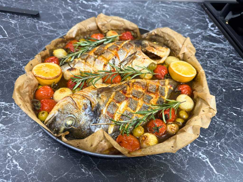 Baking dish with two roasted dorada fish, surrounded by cherry tomatoes, olives, pearl onions, lemon halves, and rosemary sprigs, placed on a black marble countertop.