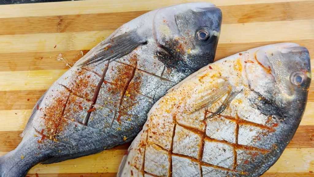Two whole dorada fish with scored skin, seasoned with spices, resting on a wooden cutting board.