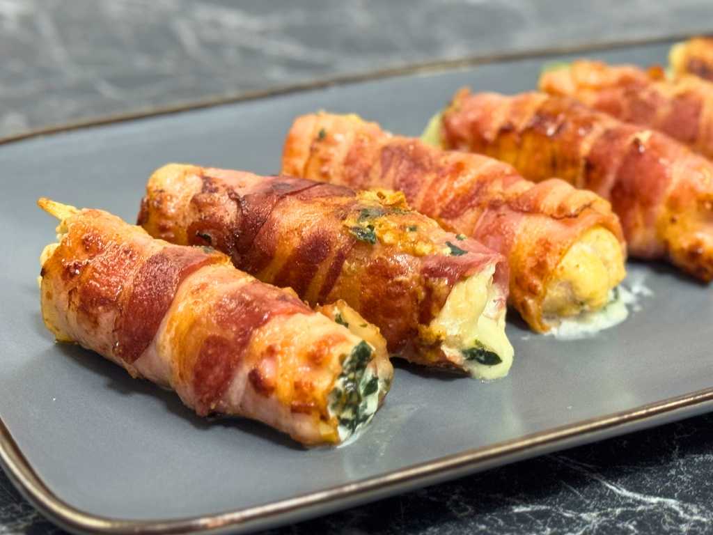 Close-up of bacon-wrapped chicken rolls stuffed with creamy spinach and cheese, served on a gray plate.
