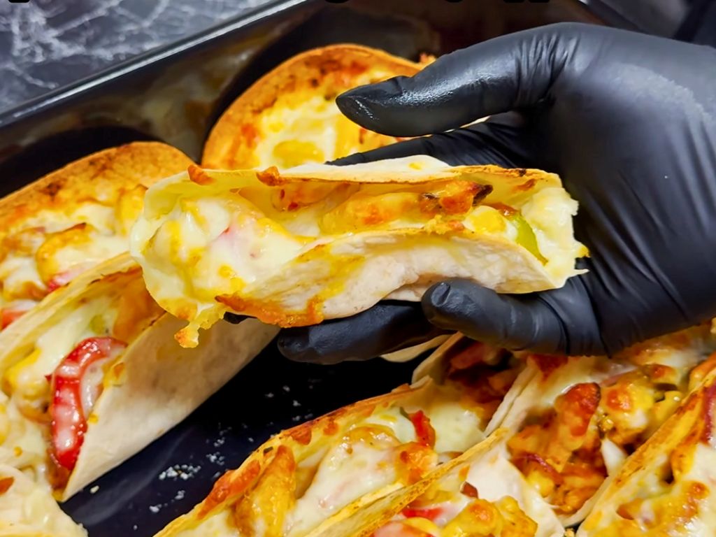 Close-up of a cheesy baked chicken taco with bell peppers and onions, held by a gloved hand over a tray of tacos.