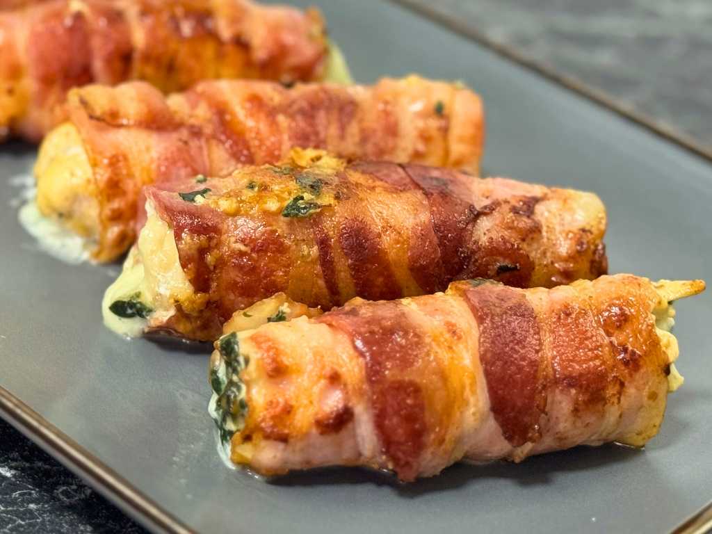Bacon-wrapped chicken rolls with golden crispy bacon and creamy spinach filling, served on a dark gray plate.