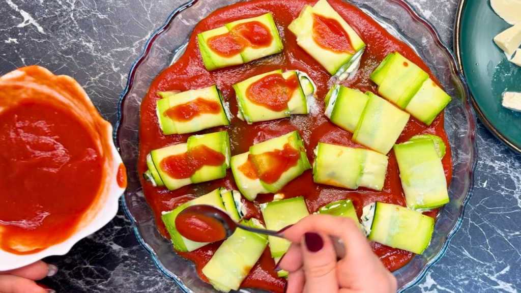 Zucchini roll-ups arranged in a glass baking dish with tomato sauce underneath, and a spoon adding more sauce on top of each roll.