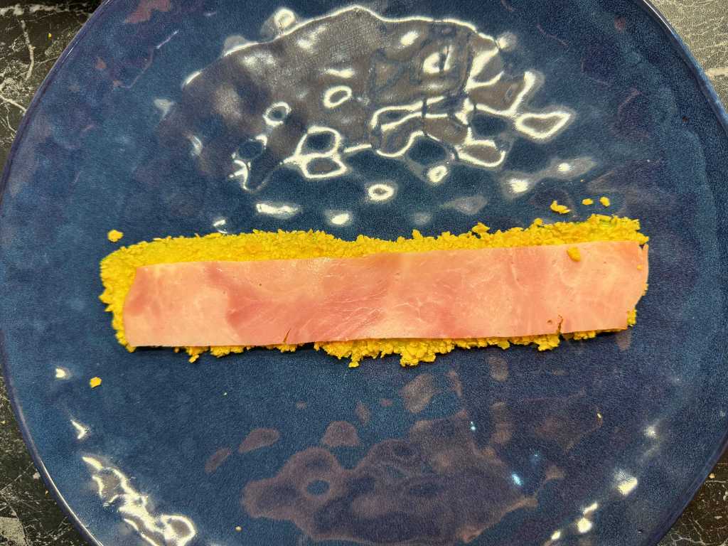 Breaded zucchini slice topped with a strip of ham, prepared for rolling into zucchini skewers with potatoes
