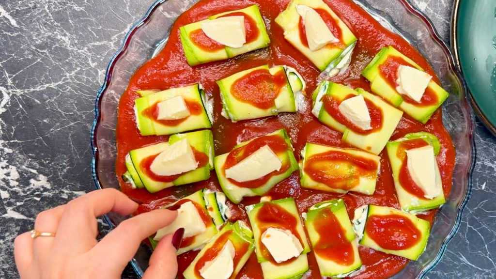 Each zucchini roll topped with a dollop of tomato sauce and a piece of mozzarella, all arranged in a glass baking dish ready for the oven.
