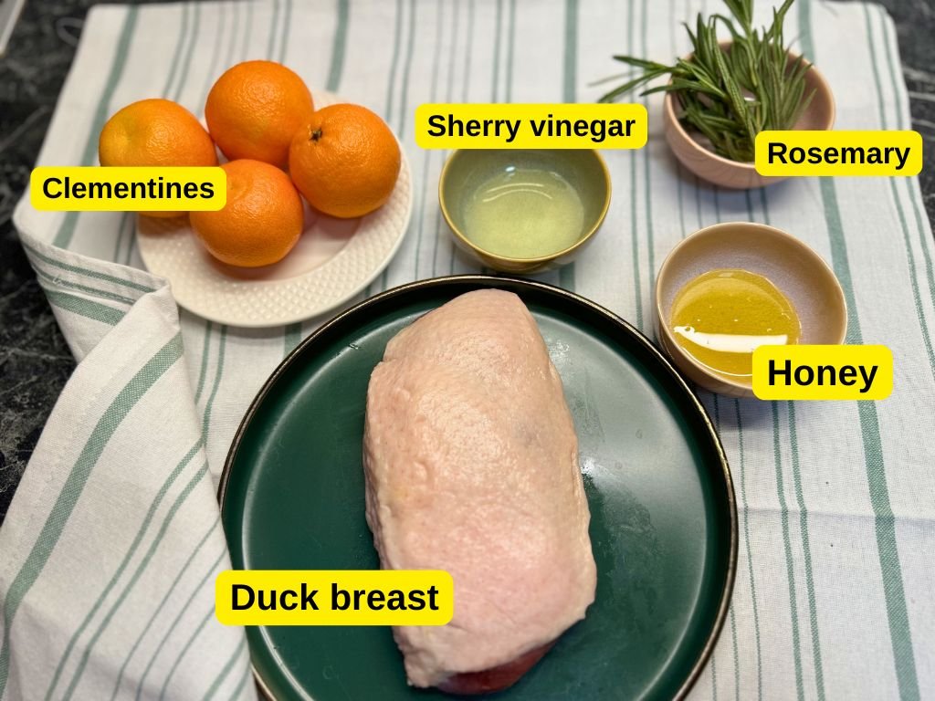 Raw duck breast on a green plate surrounded by fresh clementines, rosemary sprigs, sherry vinegar, and honey—key ingredients for a honey orange glazed duck breast recipe, with each item labeled.