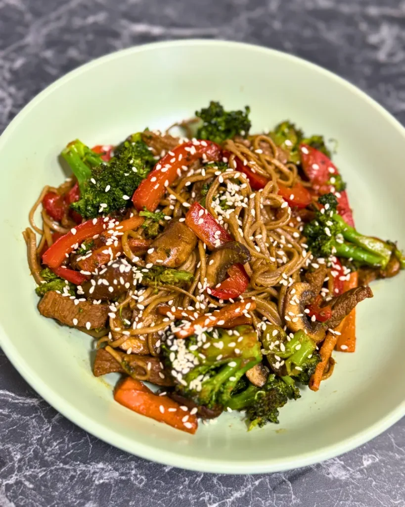Bowl of Japanese soba noodles stir fry with tender beef strips, broccoli florets, bell peppers, carrots, mushrooms, and sesame seeds on top.