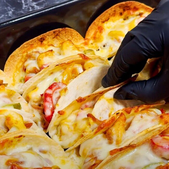 Baked chicken tacos filled with melted cheese, bell peppers, and onions, being picked up from a tray by a gloved hand.