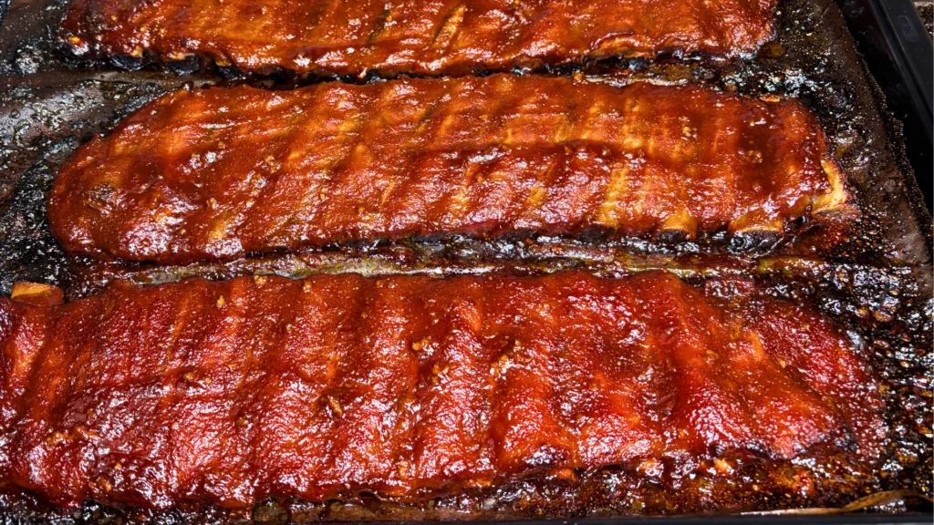 Oven-baked pork ribs coated in a sticky glaze on a dark baking tray.