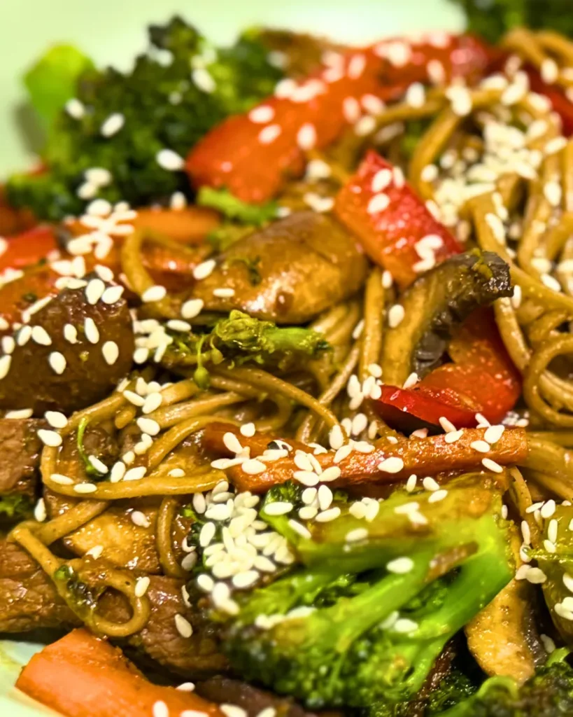 Close-up of Japanese soba noodles with sliced beef, broccoli, red bell peppers, carrots, mushrooms, and sesame seeds in a light green bowl.