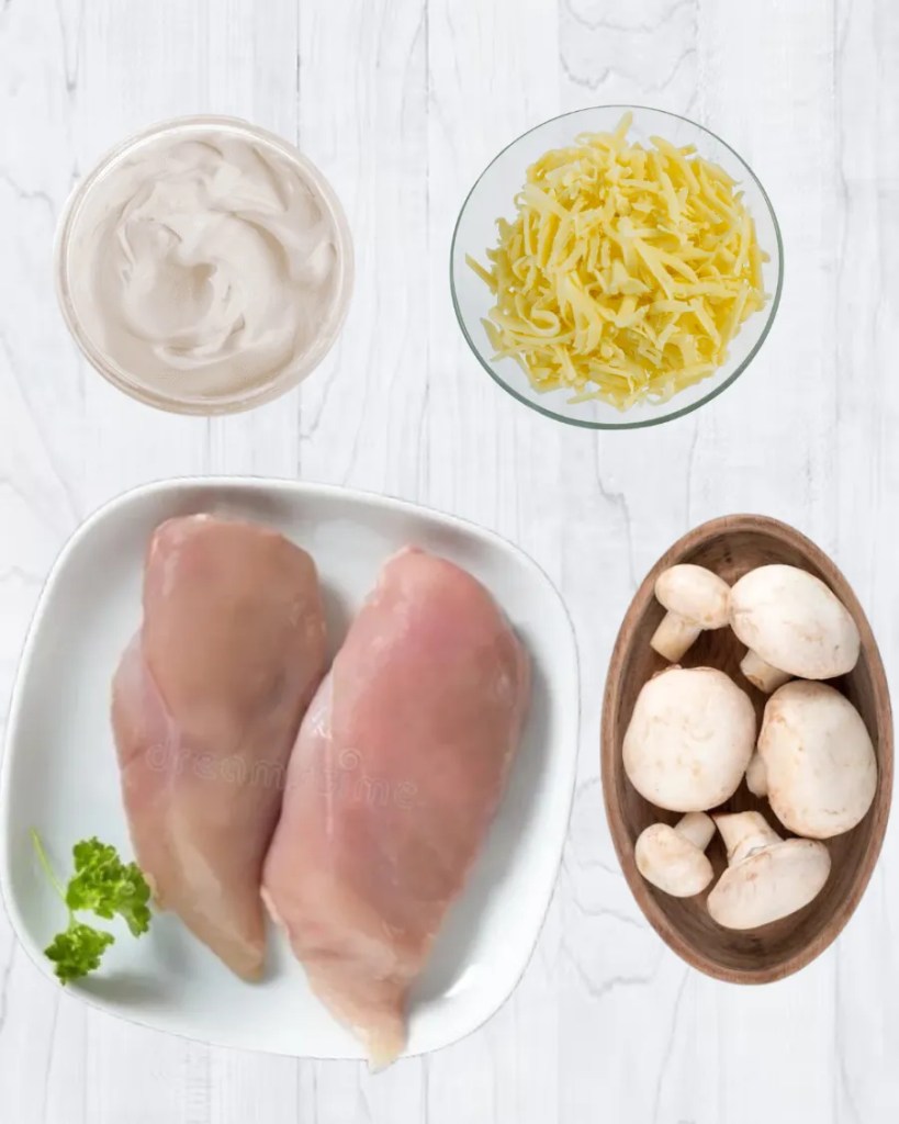 Top view of raw chicken breasts, mushrooms, grated cheese, and whipping cream arranged on a white surface, ready for cooking.