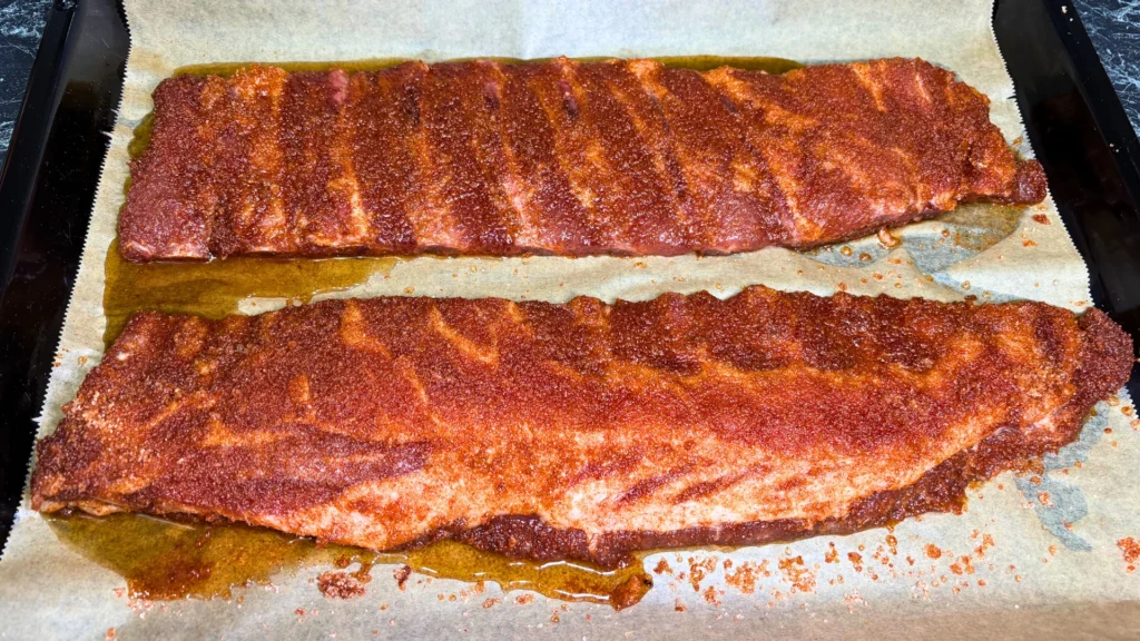 Two racks of pork ribs coated with a reddish spice rub on a parchment-lined baking tray.