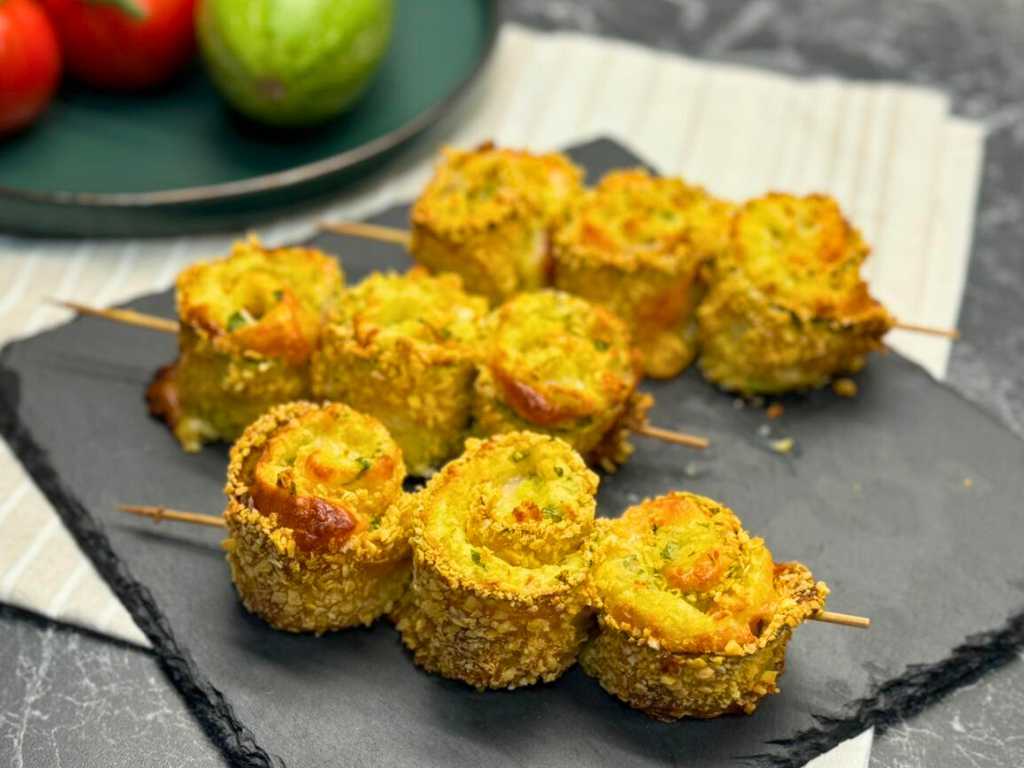 Crispy baked zucchini roll skewers with mashed potatoes, ham, and cheese, served on a slate tray with tomatoes and zucchini in the background