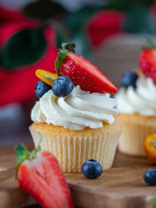 Homemade vanilla cupcakes topped with cream cheese frosting, decorated with fresh raspberries and blueberries on a white cake stand.