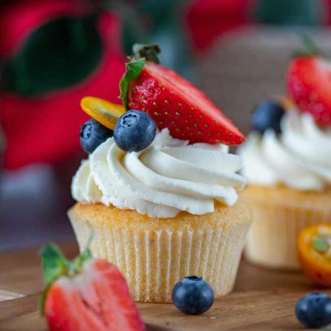 Homemade vanilla cupcakes topped with cream cheese frosting, decorated with fresh raspberries and blueberries on a white cake stand.