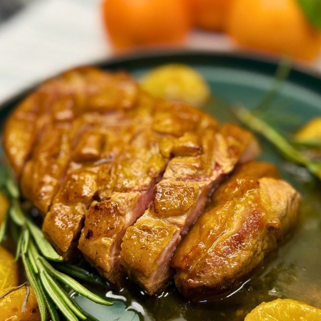 Close-up of sliced honey orange glazed duck breast with crispy golden skin and juicy pink center, served in citrus glaze with rosemary and clementines.