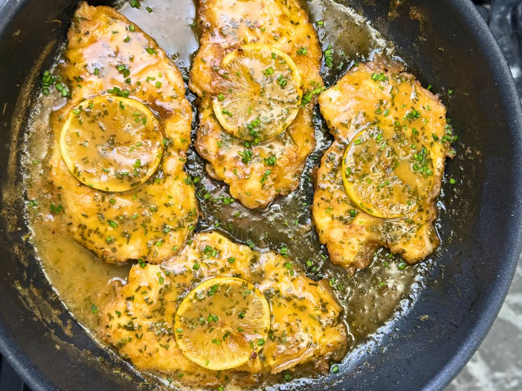 Close-up of crispy chicken cutlets simmering in a glossy lemon herb sauce, topped with lemon rounds and chopped green herbs in a frying pan.