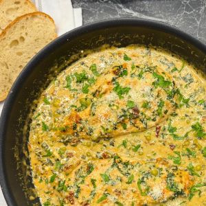 Creamy coconut milk chicken breast cooked with spinach, green peas, sun-dried tomatoes, and parmesan cheese in a black skillet, garnished with fresh parsley and served with sliced bread.