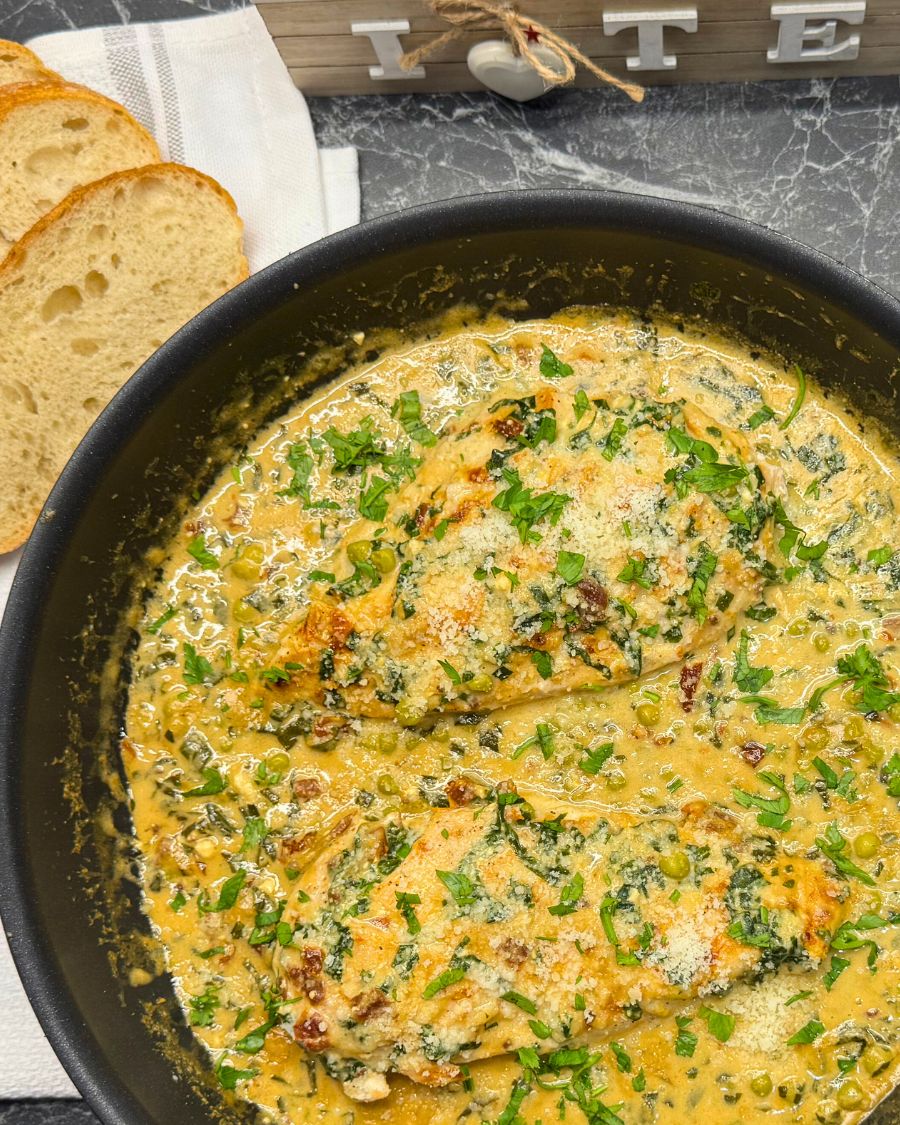 Creamy coconut milk chicken breast cooked with spinach, green peas, sun-dried tomatoes, and parmesan cheese in a black skillet, garnished with fresh parsley and served with sliced bread.