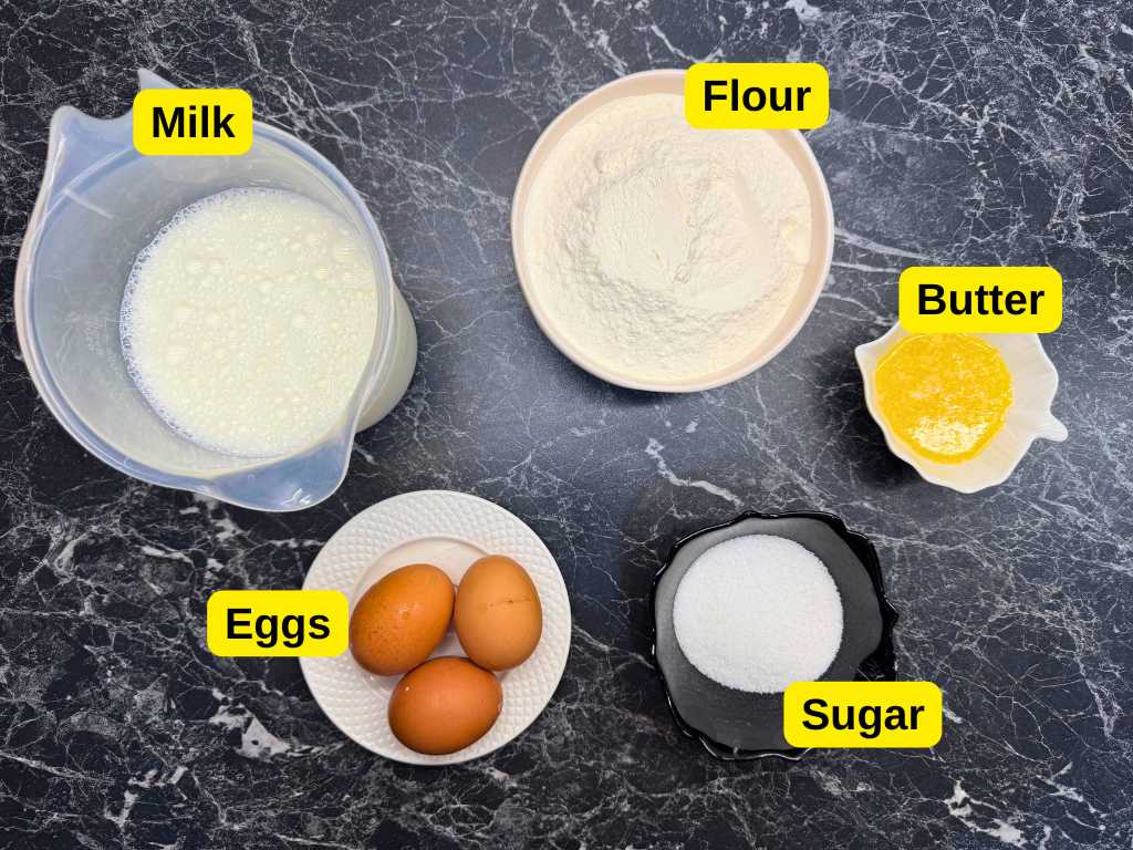 Top-down view of labeled pancake ingredients on a dark marble surface: a jug of milk, a bowl of flour, a dish of melted butter, a plate with three brown eggs, and a black bowl of granulated sugar.