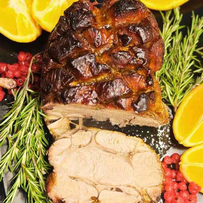 Sliced oven roasted pork neck with honey soy glaze, garnished with fresh rosemary, orange slices, and red peppercorns