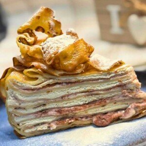 Slice of creamy pancake cake layered with whipped cream, cream cheese, and raspberry jam, topped with golden crispy pancake ribbons and dusted with powdered sugar, served on a rustic ceramic plate.