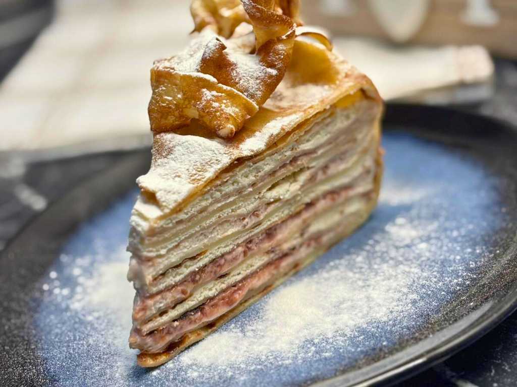 Slice of creamy layered pancake cake filled with whipped cream, cream cheese, and raspberry jam, topped with crisp pancake ribbons and dusted with powdered sugar, served on a blue ceramic plate.