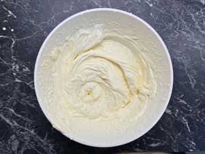 Close-up of a white mixing bowl filled with smooth, whipped cream cheese filling, swirled to a soft peak texture on a dark marble countertop.