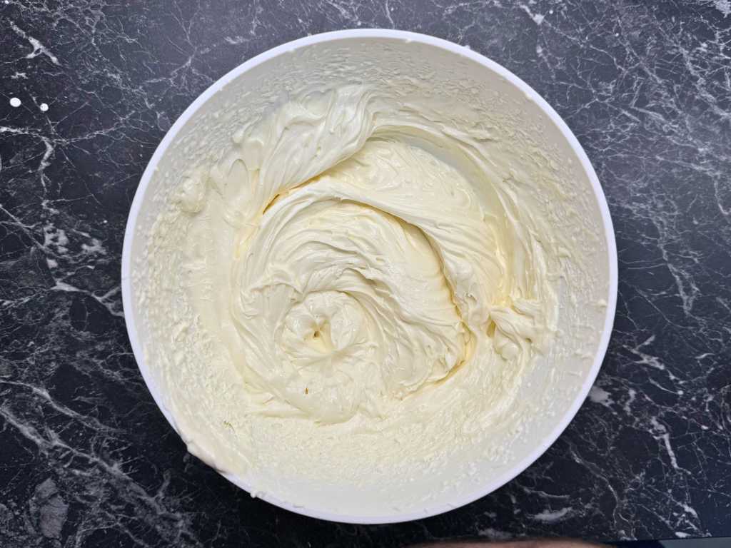 Close-up of a white mixing bowl filled with smooth, whipped cream cheese filling, swirled to a soft peak texture on a dark marble countertop.