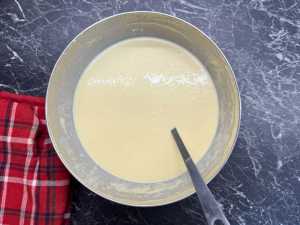 Large metal bowl filled with smooth pancake batter, resting on a dark marble surface with a red plaid kitchen towel beside it and a spoon partially immersed in the mixture.