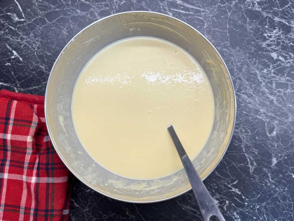 Large metal bowl filled with smooth pancake batter, resting on a dark marble surface with a red plaid kitchen towel beside it and a spoon partially immersed in the mixture.
