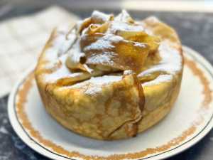 Whole creamy pancake cake wrapped in golden crepes and tied at the top with crispy pancake ribbons, dusted generously with powdered sugar and served on a round plate.