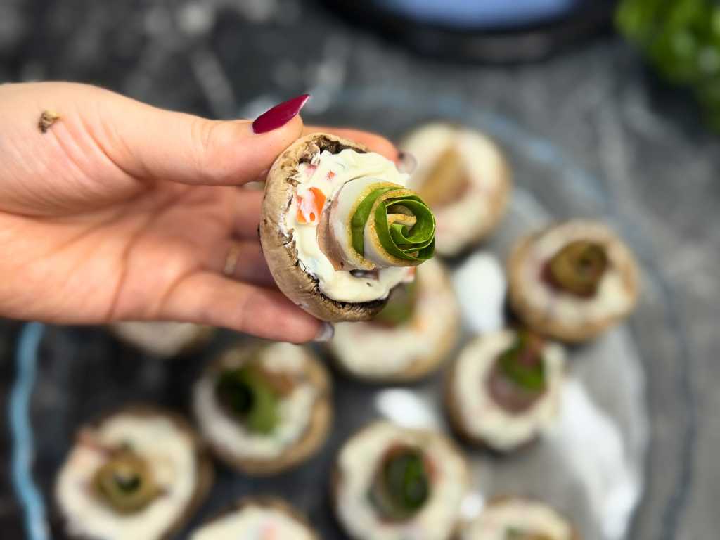 Hand holding a stuffed mushroom topped with a jamón and spinach roll, showing the assembly step before baking.