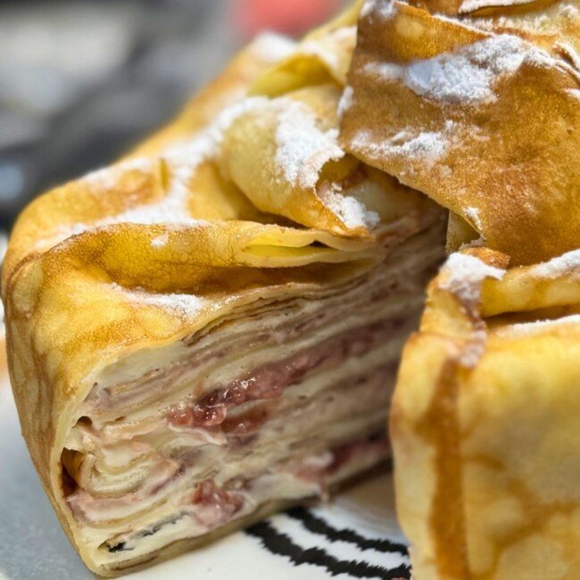 Close-up of a sliced creamy pancake cake showing delicate crepe layers filled with cream cheese, whipped cream, and raspberry jam, wrapped in golden pancakes and dusted with powdered sugar.